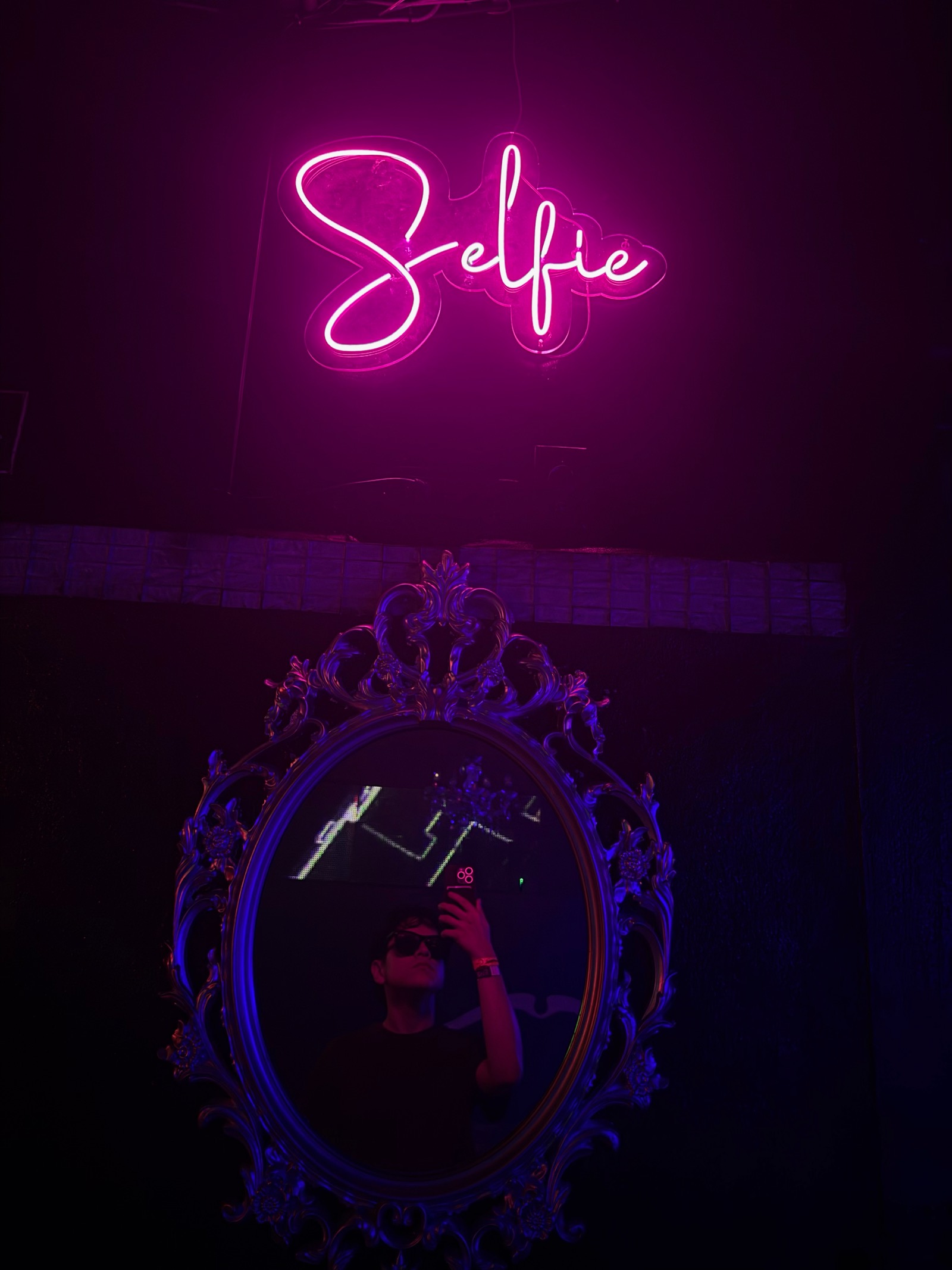 Letrero de neón rosado que dice “Selfie” sobre un espejo ornamentado.