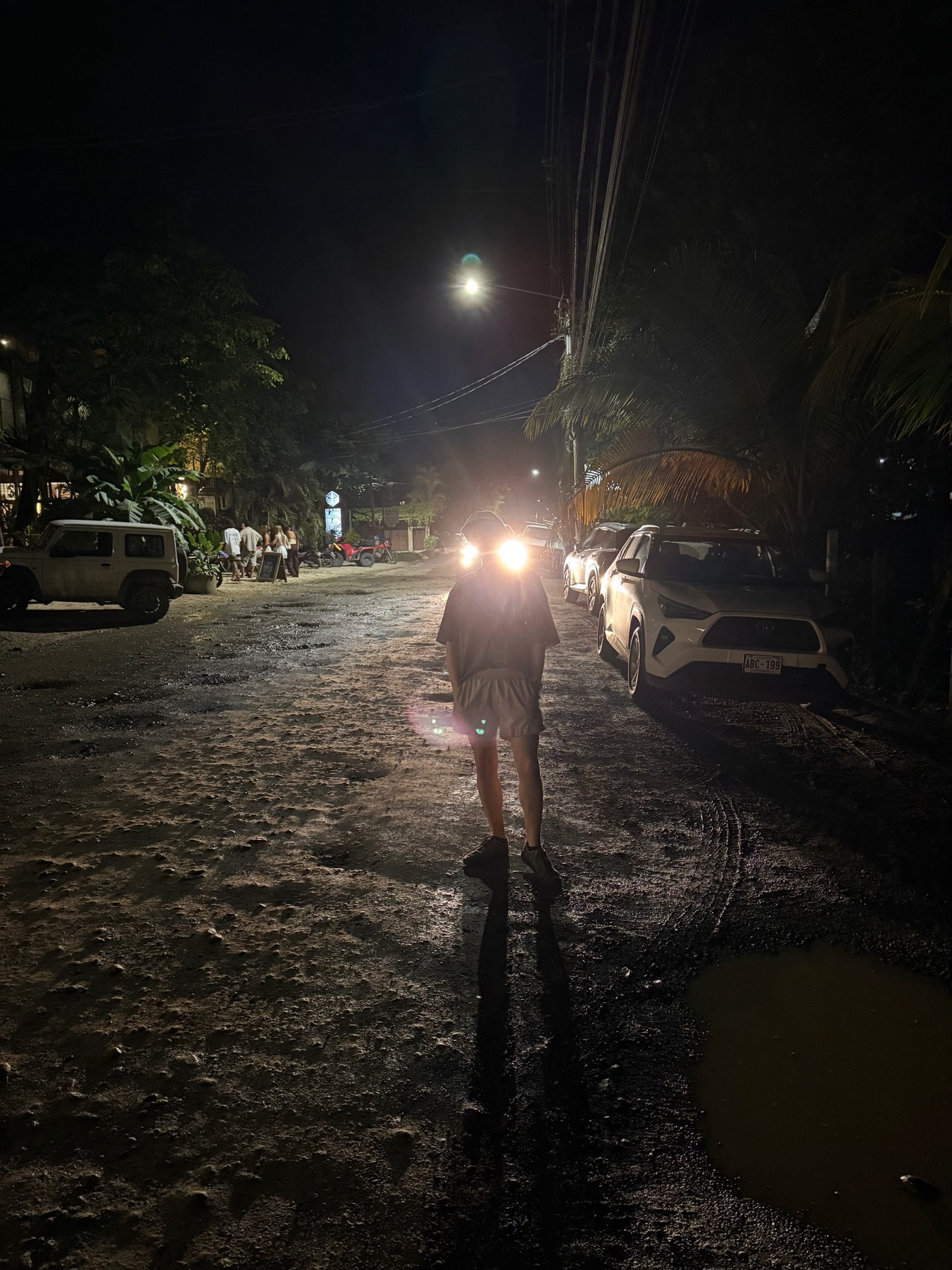 Persona caminando por una calle oscura hacia las luces de varios vehículos.
